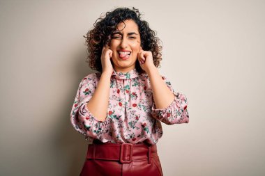 Genç, kıvırcık Arap bir kadın, çiçekli tişört giyiyor. İzole edilmiş beyaz arka plan üzerinde duruyor. Yüksek sesli müziğin gürültüsü yüzünden rahatsız edici ifadeleri olan kulaklarla kulakları örtüyor. Sağır kavramı.