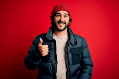 Sakallı, sıradan bir ceket giyen ve kırmızı arka planda yün şapka takan genç yakışıklı bir adam elinle mutlu bir jest yapıyor. Kameraya bakıp başarıyı gösteren ifade onaylanıyor.