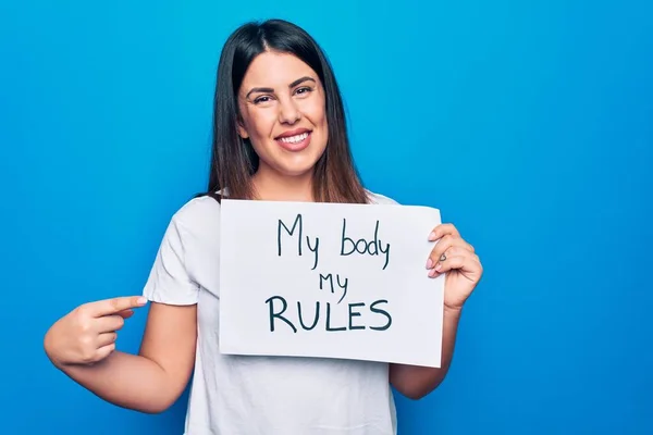 Young beautiful woman asking for women rights holding paper with my body my rules message smiling happy pointing with hand and finger