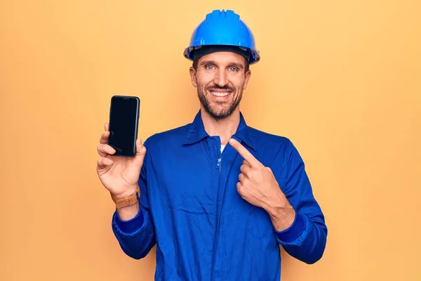 制服を着た若いハンサムな労働者の男とハード帽子を保持スマートフォンショー画面笑顔幸せなポインティングで手と指 — ストック写真