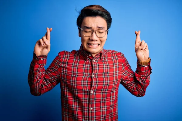 青い背景にカジュアルなシャツと眼鏡を身に着けている若いハンサムな中国人男性ジェスチャー指は希望と目を閉じて笑顔を越えました 幸運と迷信の概念 — ストック写真