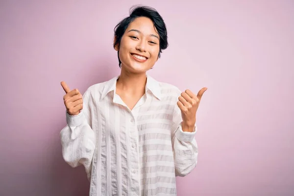 孤立したピンクの背景の成功の印の上に立つカジュアルシャツを身に着けている若い美しいアジアの女の子は手で肯定的なジェスチャーをする 親指を笑顔と幸せ 陽気な表情と勝者のジェスチャー — ストック写真
