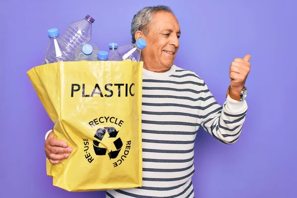 高齢者のリサイクル袋でペットボトルをリサイクルし 紫色の背景を指し 幸せな顔を笑顔で親指で横に示す — ストック写真