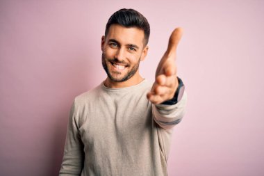 İzole edilmiş pembe arka planda duran sıradan bir süveter giyen yakışıklı genç adam dostça bir selamlama ve hoş geldin hediyesi olarak el sıkışma sunuyor. Başarılı bir iş..