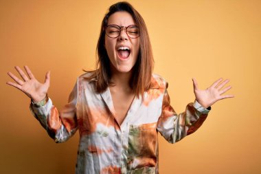 Genç ve güzel esmer kız gündelik gömlek ve gözlük takarak izole edilmiş sarı arka planda başarı için çılgın ve çılgın kutlamalar yapıyor kollarını kaldırmış ve gözlerini kapatmış heyecanlı çığlıklar atıyor. Kazanan konsept