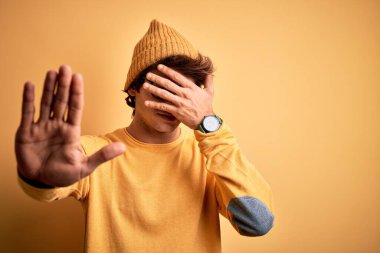 Genç, yakışıklı bir adam günlük tişört ve gözlük takıyor. İzole edilmiş sarı arka plan, gözleri ellerle kaplıyor ve üzgün ve korku ifadeleriyle jest yapıyor. Utanç verici ve olumsuz bir kavram..