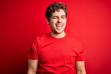 Kıvırcık saçlı, kırmızı arka planda sıradan tişörtler giyen genç sarışın yakışıklı adam kameraya seksi bir ifadeyle bakıyor, neşeli ve mutlu bir yüzle..