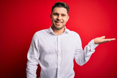 Mavi gözlü, zarif gömlekli genç bir iş adamı kırmızı izole edilmiş arka planda dikiliyor neşeli bir şekilde gülümsüyor ve avuç içi kameraya bakıyor..