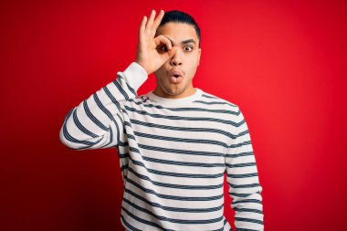 İzole edilmiş kırmızı arka planda dikilen sıradan çizgili tişört giyen genç Brezilyalı adam şaşırmış yüz ifadesiyle şaşırmış, parmaklarından bakan gözler. İnançsız ifade.
