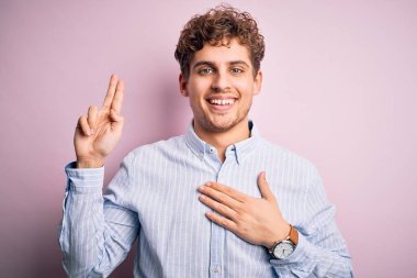 Sarışın, kıvırcık saçlı, çizgili tişörtlü, beyaz arka planda gülümseyen, elleri göğsünde ve parmakları havada, sadakat yemini eden genç bir adam.