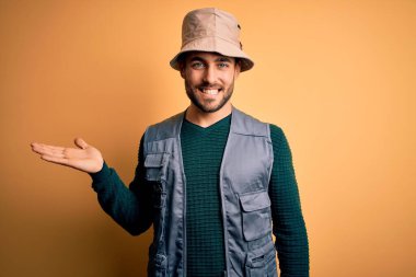 Tatilde sakallı yakışıklı turist sarı arka planda kaşif şapkası takıyor neşeli bir şekilde sunuyor ve avuç içi kameraya bakıyor..