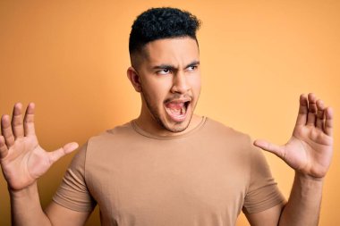 Yalnız sarı arka planda dikilen rahat tişörtlü genç ve yakışıklı bir adam sinirli bir şekilde bağırıyor ve agresif bir ifadeyle kollarını kaldırıyor. Hayal kırıklığı konsepti.