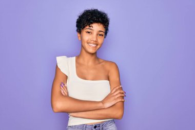 Genç ve güzel Afro-Amerikan kadın mor arka planda rahat bir tişört giyiyor ve kollarını kavuşturup kameraya bakıyor. Pozitif kişi.