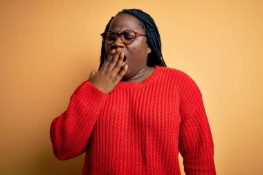 Sarı arka planda sıradan süveter giyen Afrikalı Amerikalı artı cüsseli kadın ağzıyla esnemekten sıkılmış. Huzursuz ve uykulu..