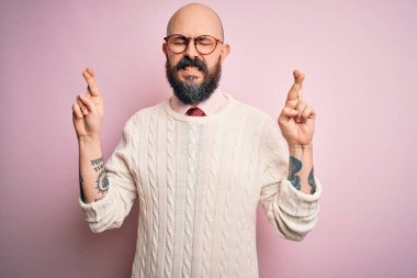 Sakallı, dövmeli, gözlüklü, pembe arka planda süveter giyen yakışıklı kel adam. Umut dolu ve gözleri kapalı bir şekilde gülümseyerek işaret ediyor. Şans ve batıl inanç..
