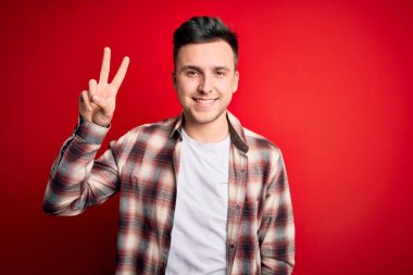 Kırmızı izole edilmiş arka plan üzerinde gündelik modern gömlekler giyen yakışıklı beyaz adam kameraya bakıp zafer işareti yapıyor. İki numara..