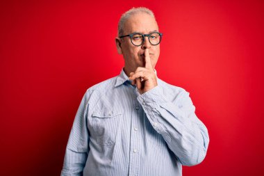 Orta yaşlı, yakışıklı, beyaz, sıradan çizgili tişört giyen ve kırmızı arka planda gözlük takan bir adam sessiz olmayı diliyor. Sessizlik ve gizli konsept.