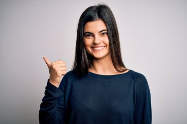 Genç, güzel, esmer, sıradan bir süveter giyen, beyaz arka planda duran mutlu yüzüyle gülümseyen ve baş parmağıyla yan tarafı gösteren bir kadın..