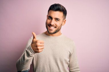 Soyutlanmış pembe arka plan üzerinde duran sıradan süveter giyen genç yakışıklı adam mutlu bir el hareketi yapıyor. Kameraya bakıp başarıyı gösteren ifade onaylanıyor.