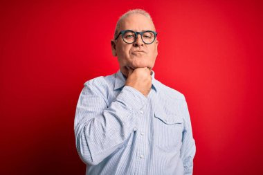 Orta yaşlı yakışıklı beyaz bir adam kırmızı arka planda günlük çizgili gömlek ve gözlük takıyor. Ağrılı boynuna dokunuyor. Grip için boğaz ağrısı, mantar ve enfeksiyon kapmış.