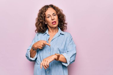 Orta yaşlı güzel bir kadın pembe arka planda dikilmiş sıradan bir kot gömlek giyiyor. Aceleyle saati, sabırsızlığı ve gecikmeyi işaret ediyor.