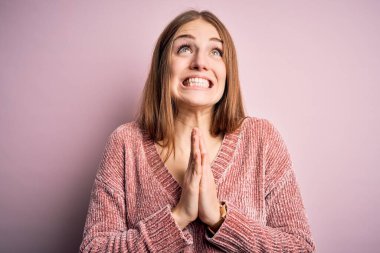 Genç, güzel, kızıl saçlı bir kadın pembe arka plan üzerine günlük süveter giyip elleri bağlı yalvarıyor ve dua ediyor. Yüzünde umut ifadesi var. Çok duygusal ve endişeli. Yalvarıyorum..