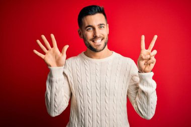 İzole edilmiş kırmızı arka plan üzerinde duran, rahat beyaz süveter giyen genç yakışıklı adam yedinci parmaklarıyla işaret ederken kendinden emin ve mutlu bir şekilde gülümsüyor..