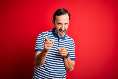 Orta yaşlı, beyaz, sıradan çizgili polo giyen, izole edilmiş kırmızı arka planda durup mutlu ve komik yüzlü parmakları kameraya gösteren bir adam. İyi enerji ve titreşimler.