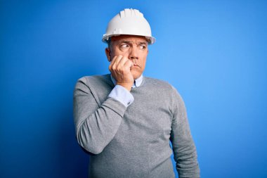 Mavi arka planda güvenlik kaskı takan orta yaşlı, yakışıklı, gri saçlı mühendis elleri ağızda tırnak yerken stresli ve gergin görünüyor. Endişe sorunu.