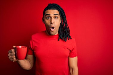 Genç Afrikalı Afro Amerikalı adam kırmızı arka planda bir fincan kahve içiyor. Şaşırmış bir yüzle korkmuş ve korku ifadesinden heyecanlanmış.