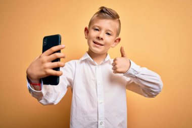 Küçük beyaz bir çocuk akıllı telefon kullanıyor ve büyük bir gülümsemeyle telefonun ekranına bakıyor. Tamam işareti yapıyor, parmaklarıyla başparmağını kaldırıyor, mükemmel bir işaret.