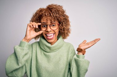 Afro-Amerikan optik kadın, kıvırcık saçlı, gözlüklü, izole edilmiş beyaz arka planda mutlu ve heyecanlı, kazanan yüz ifadesi zaferi büyük bir gülümsemeyle kutluyor ve ellerini kaldırıyor.