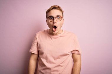 Genç, yakışıklı, günlük tişört giyen, pembe arka planda duran, korku dolu, şaşkınlık ve şaşkınlık dolu ifade, korku ve heyecan dolu yüz ifadesinden korkan genç bir adam..