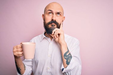 Sakallı, dövmeli, pembe arka planda kahve içen yakışıklı kel bir adam.