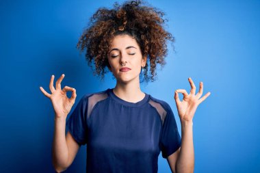 Kıvırcık saçlı ve piercing giyen, sıradan mavi tişörtlü, gözleri kapalı, meditasyon yaparken parmaklarıyla gülümseyen güzel bir kadın. Yoga konsepti.