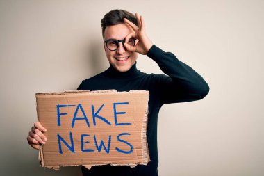 Genç yakışıklı beyaz adam yanlış bilgilendirme için protesto kartı tutuyor ve gülen yüzlü sahte haberler veriyor. Elle gözün içine bakarak işaret veriyor.