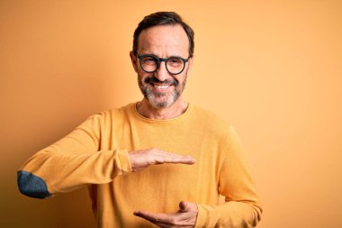 Orta yaşlı, beyaz bir adam, günlük süveter ve gözlük takıyor, sarı arka planda el kol hareketleri yapıyor, büyük ve büyük boyut işareti, ölçü sembolü. Kameraya bakıp gülümsüyor. Ölçüm konsepti.