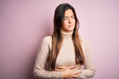 Balıkçı yaka kazak giyen genç ve güzel bir kız midesi ağrıyan, hazımsızlık çeken, acı veren bir hastalıktan dolayı izole edilmiş pembe arka planda duruyor. Ache kavramı.