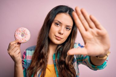 Genç, güzel esmer kadın izole edilmiş arka planda tatlı pembe çörekler yiyor. Açık elle dur işareti yapıyor. Ciddi ve kendinden emin bir ifadeyle, savunma hareketi yapıyor.