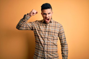 Genç ve yakışıklı bir adam, izole edilmiş sarı arka planda dikiliyor kızgın ve kızgın bir şekilde yumruğunu kaldırıyor öfkeyle bağırırken. Öfke ve agresif kavram.