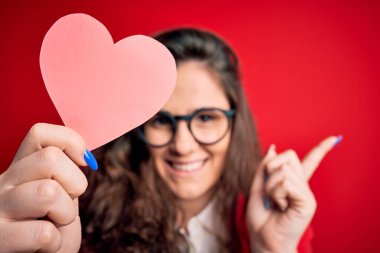 Kıvırcık saçlı genç bir kadın izole edilmiş kırmızı arka planda kağıt bir kalple elini ve parmağını kenarı işaret ederek çok mutlu oluyor.