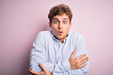 Sarışın, kıvırcık saçlı, beyaz arka planda çizgili gömlek giyen, kışın soğuktan titreyen ve donan, yüzünde üzgün ve şok ifadesi olan genç bir adam.