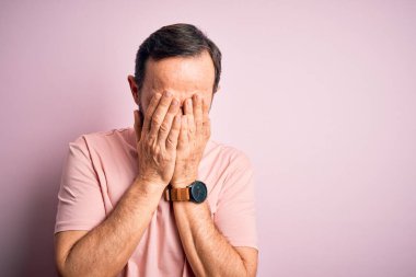 Orta yaşlı, ağlak, günlük tişört giyen, pembe arka planda dikilen, ağlarken yüzünü ellerle kapatan bir adam. Depresyon kavramı.