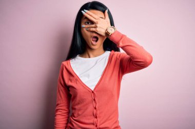 Genç, güzel, Çinli bir kadın pembe arka plan üzerine sıradan bir süveter giyer. Şoka girer, yüzünü ve gözlerini kapar ve utanmış bir ifadeyle parmaklarından bakar..