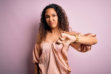 Kıvırcık saçlı, pembe arka planda sıradan tişörtler giyen, mutsuz, kızgın, reddedilmiş ve olumsuz jestli genç bir kadın. Kötü ifade.