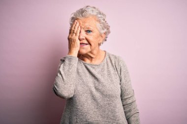 Son sınıf güzeli, günlük tişört giyen, pembe arka planda duran, bir gözünü elinle kapayan, yüzünde kendine güvenen bir gülümseme ve sürpriz duygular taşıyan bir kadın..