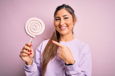 Genç, güzel esmer kadın izole edilmiş pembe arka planda şeker yiyor. Elinle ve parmağınla işaret ederek çok mutlu.