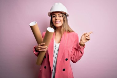 Genç ve güzel esmer mimar kadın kaskını takmış elinde planları tutarken mutlu bir şekilde elini ve parmağını kenarı işaret ediyor.