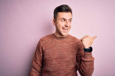 Pembe izole edilmiş arka planda sıradan bir kazak giyen yakışıklı beyaz adam gülümsüyor ve başparmağıyla yana bakıyor..
