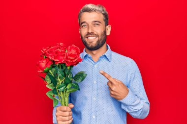 Genç, sarışın, romantik bir adam kırmızı arka planda gül buketi tutuyor. Mutlu bir şekilde elini ve parmağını işaret ediyor.